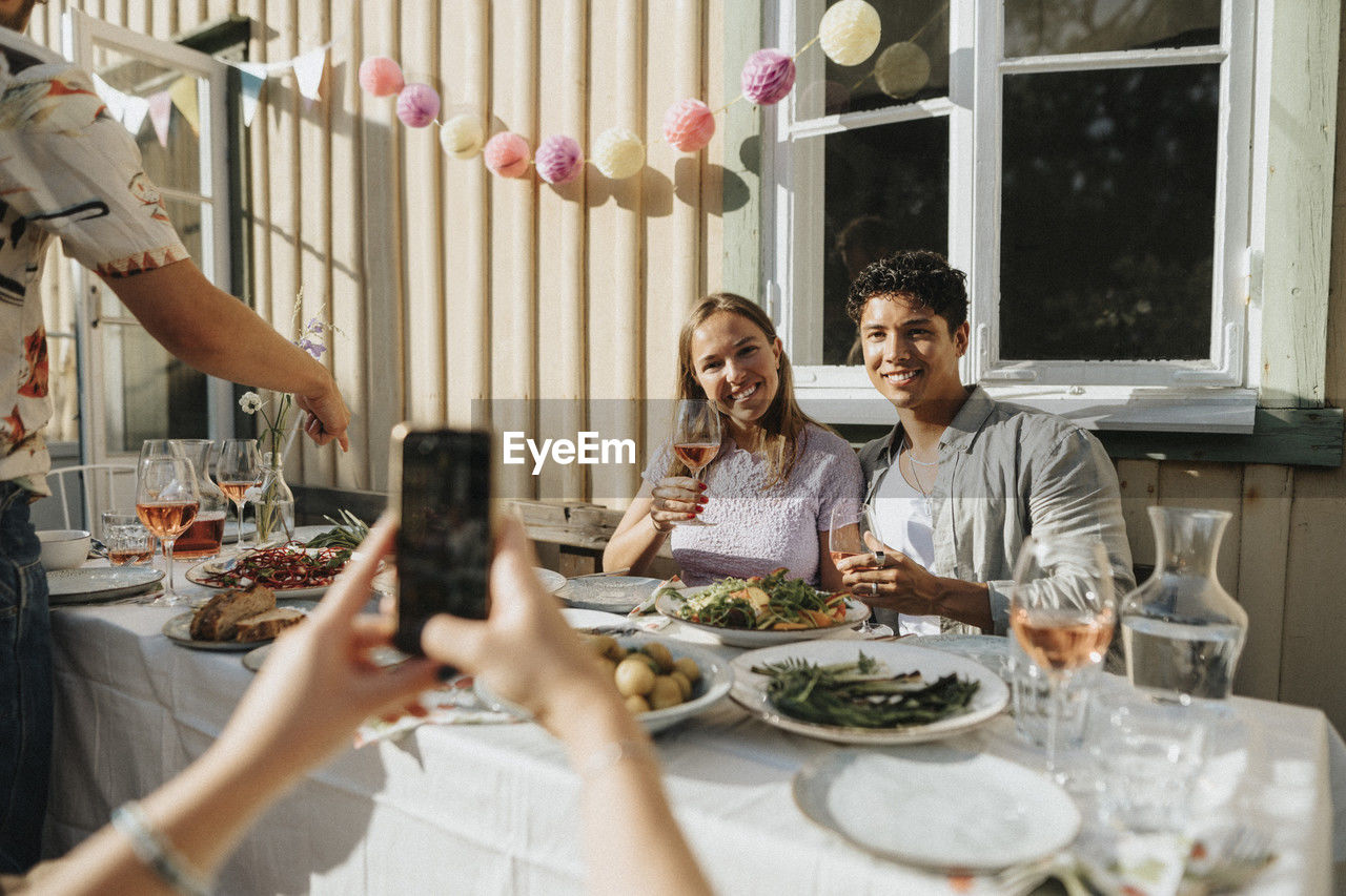 Woman clicking picture of male and female friends holding wineglass during dinner party at cafe