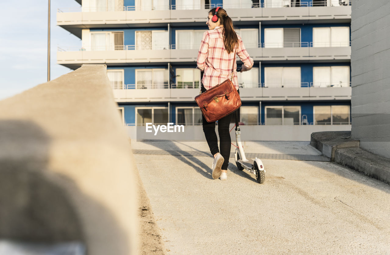 Young woman with headphones pushing electric scooter on parking deck