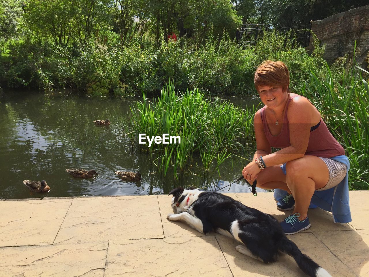 Woman crouching with dog on retaining wall by lake on sunny day