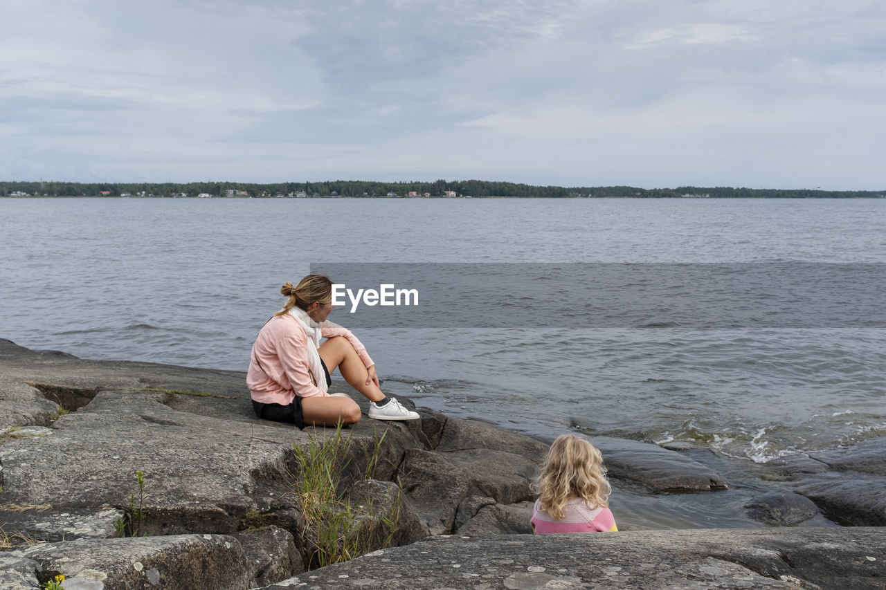 Woman and young girl sitting on rock by sea against sky