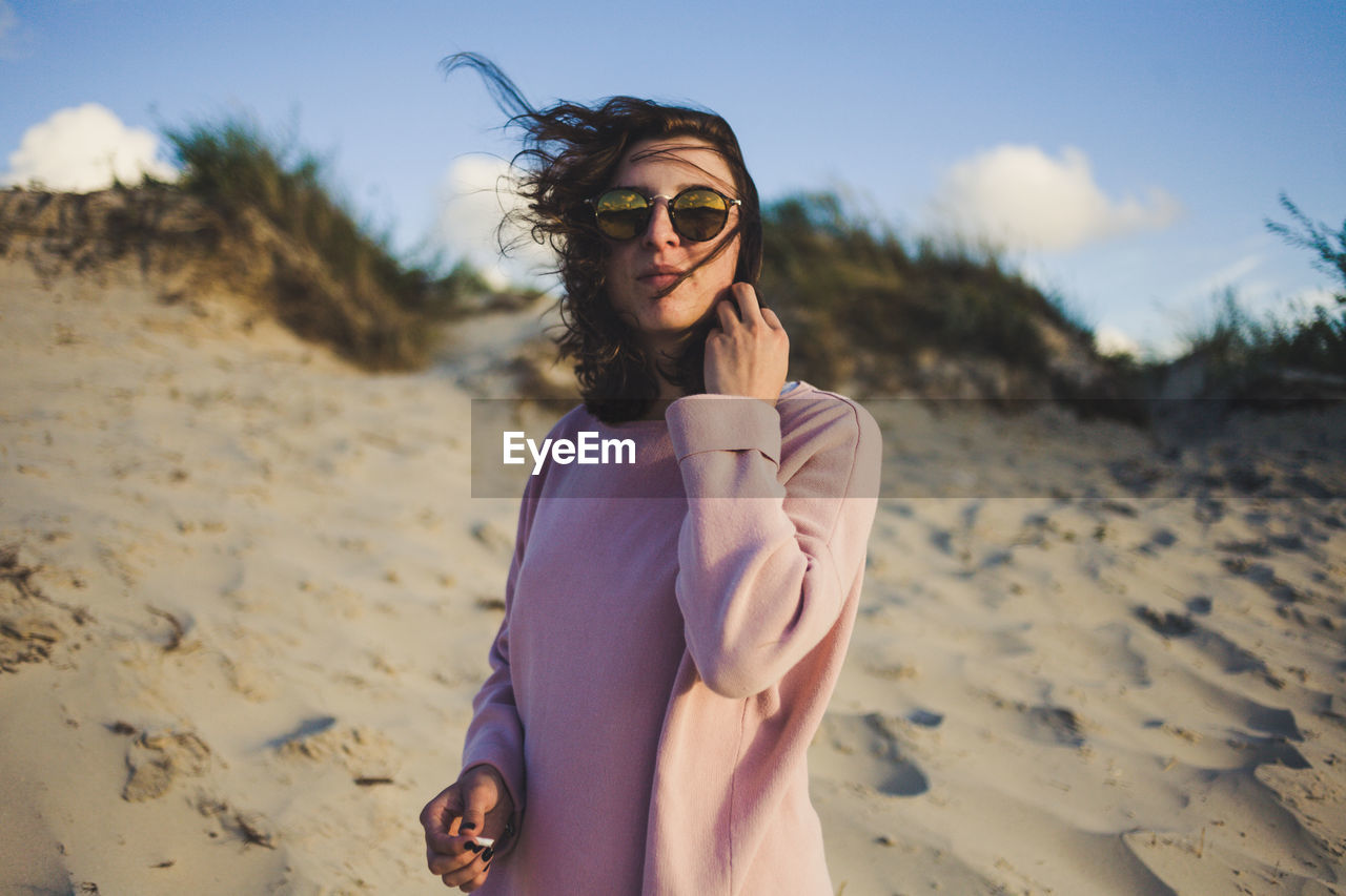 Young woman wearing sunglasses while standing at beach against sky