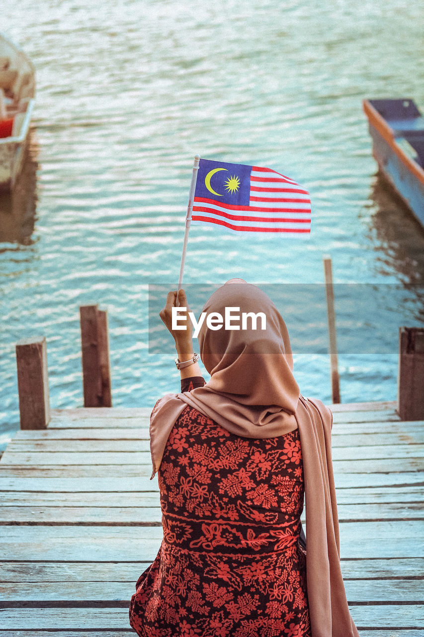 Rear view of woman wearing hijab holding malaysian flag sitting on pier by lake