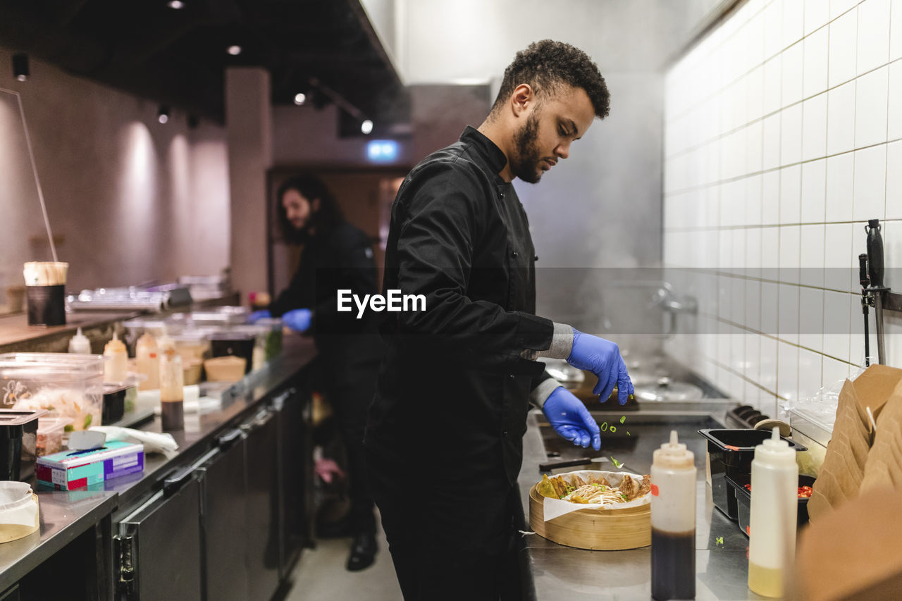 Male waiter sprinkling scallion on dumplings in bowl over kitchen counter at restaurant