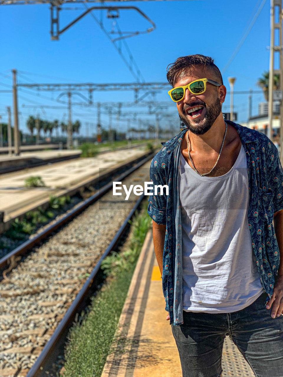 Smiling man wearing sunglasses standing on railroad platform against sky