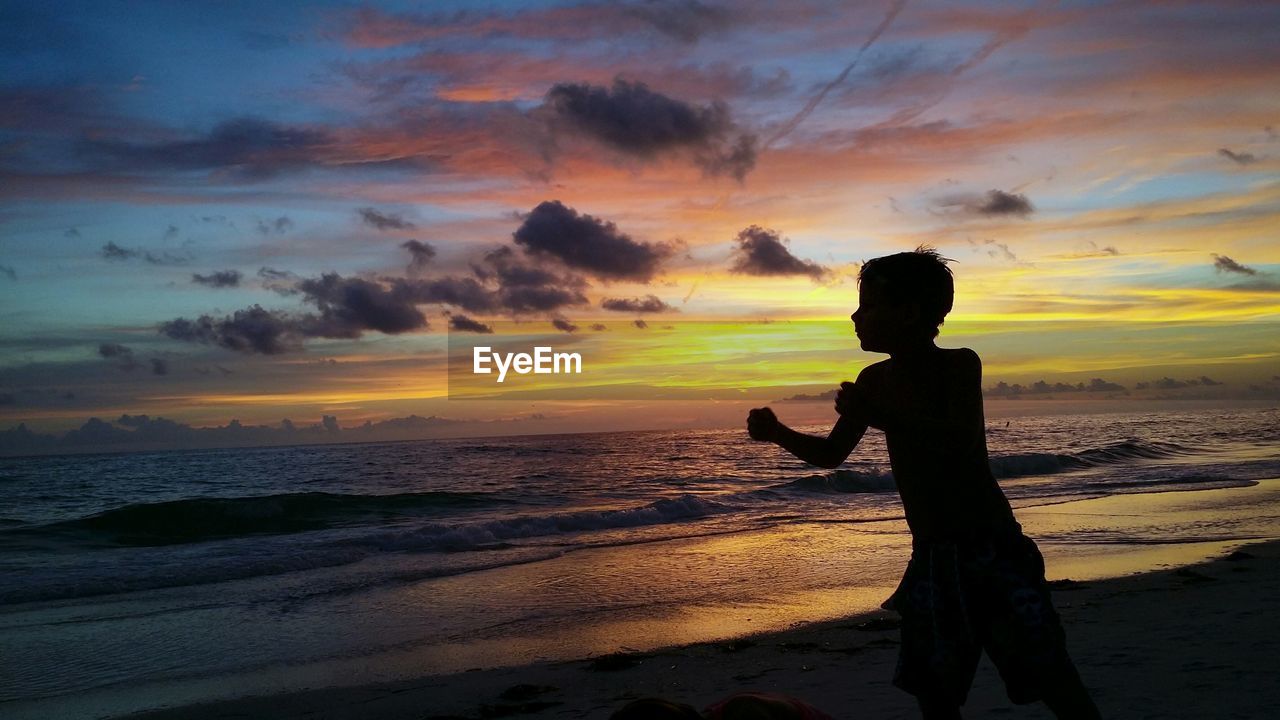 Silhouette boy on beach against orange sky