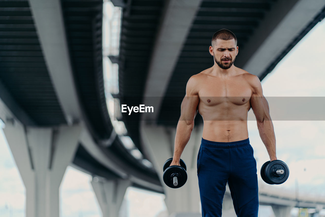 Shirtless young man lifting dumbbells while standing outdoors