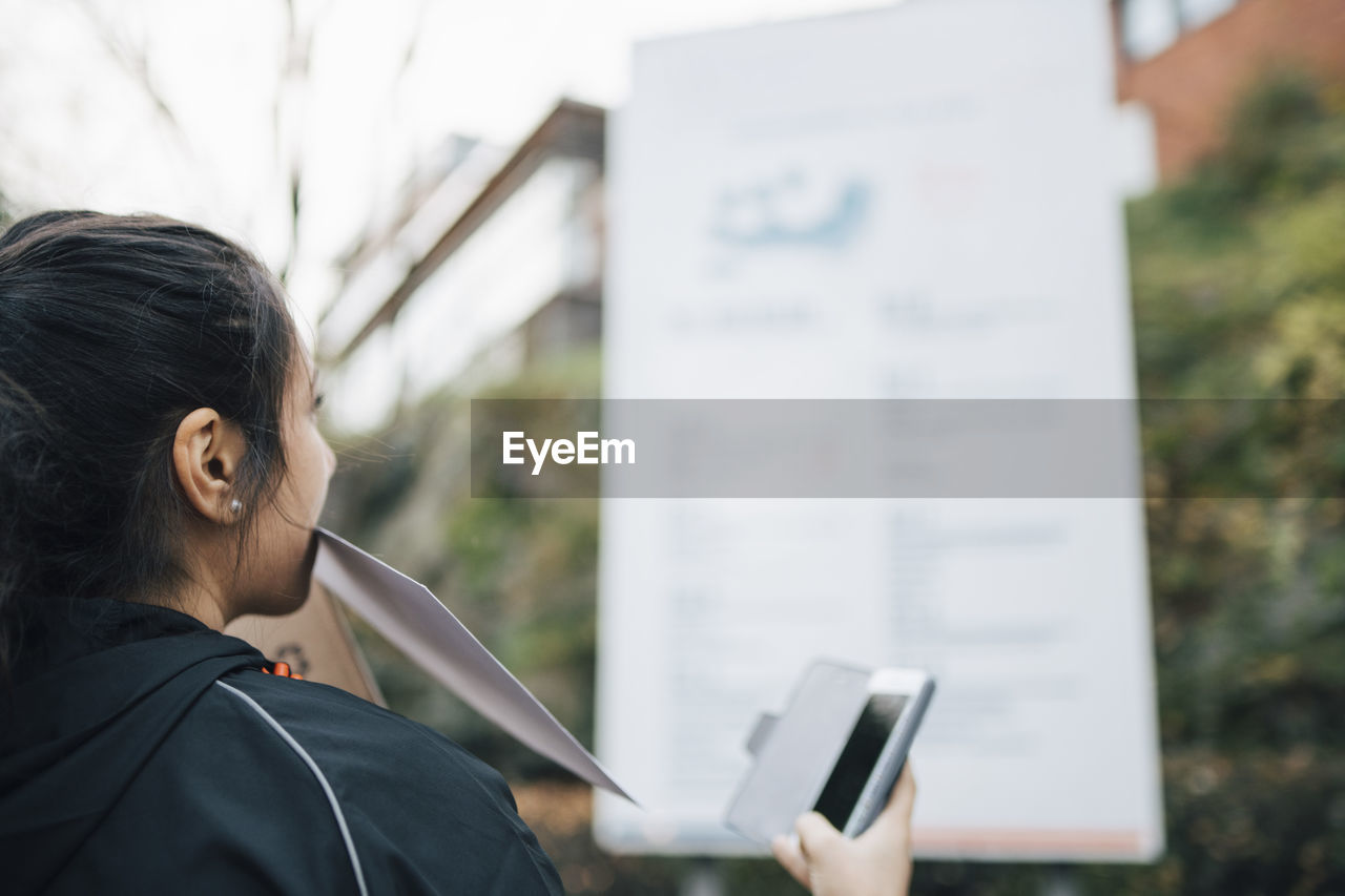 Female messenger carrying document in mouth and holding smart phone looking at billboard