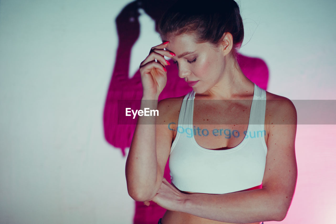 YOUNG WOMAN LOOKING AT CAMERA WHILE STANDING AGAINST WALL