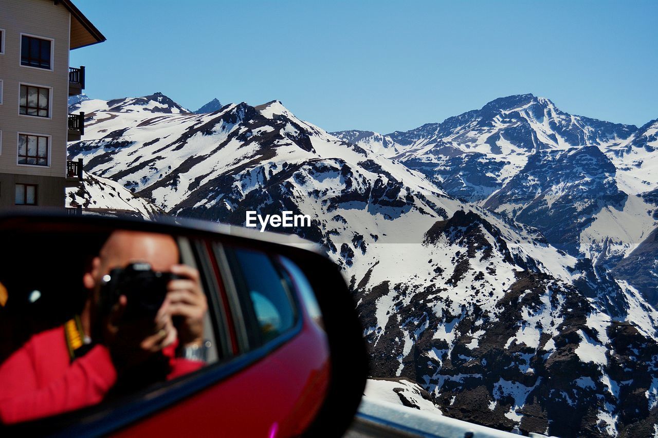 Reflection of man on side-view mirror of car against snowcapped mountain