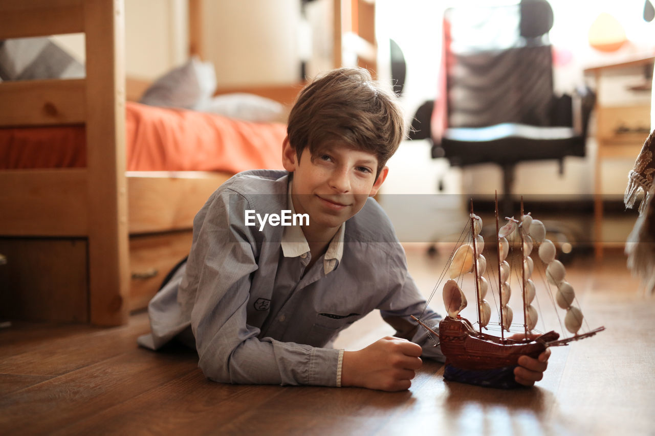 Boy teen plays with ship model on the floor in real children's room