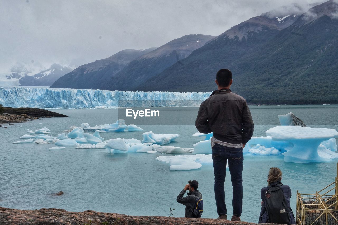 Rear view of people by lake against mountain and sky during winter