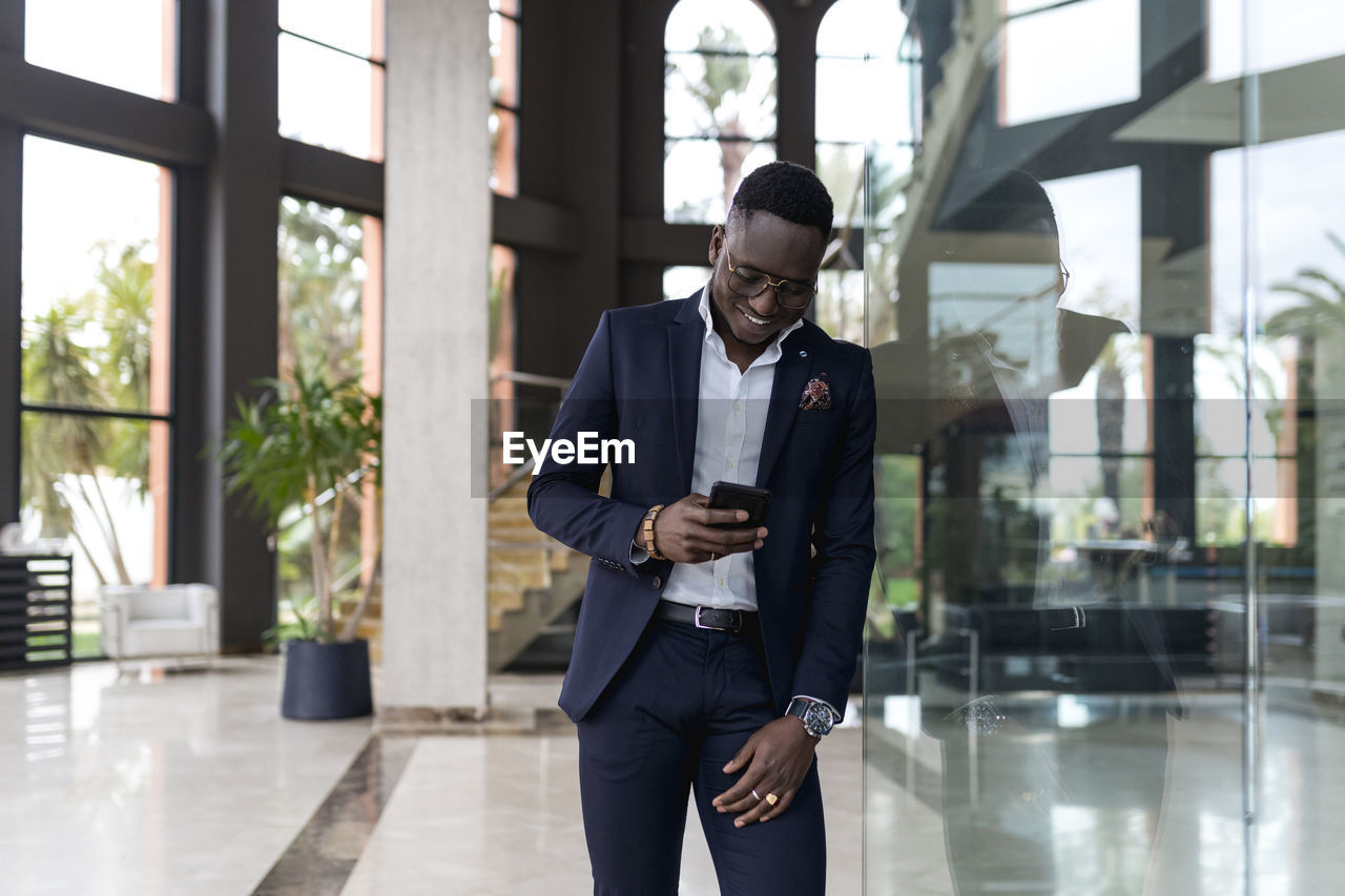 Smiling male entrepreneur using smart phone while leaning on glass wall in hotel lobby