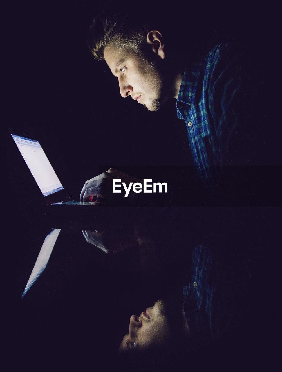 Reflection of young man on glass while using laptop in darkroom