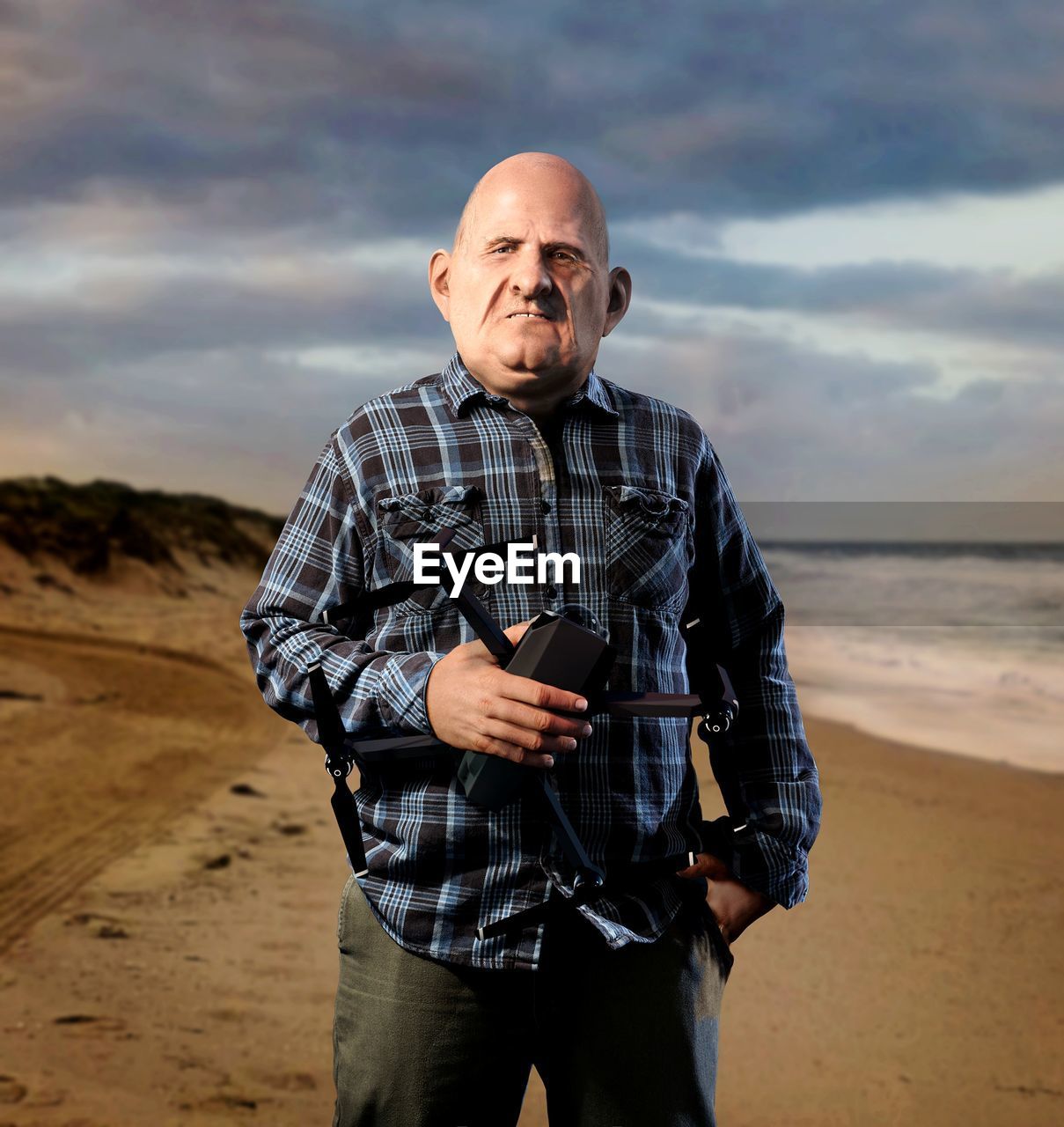 Portrait of mature man with camera standing at beach against cloudy sky