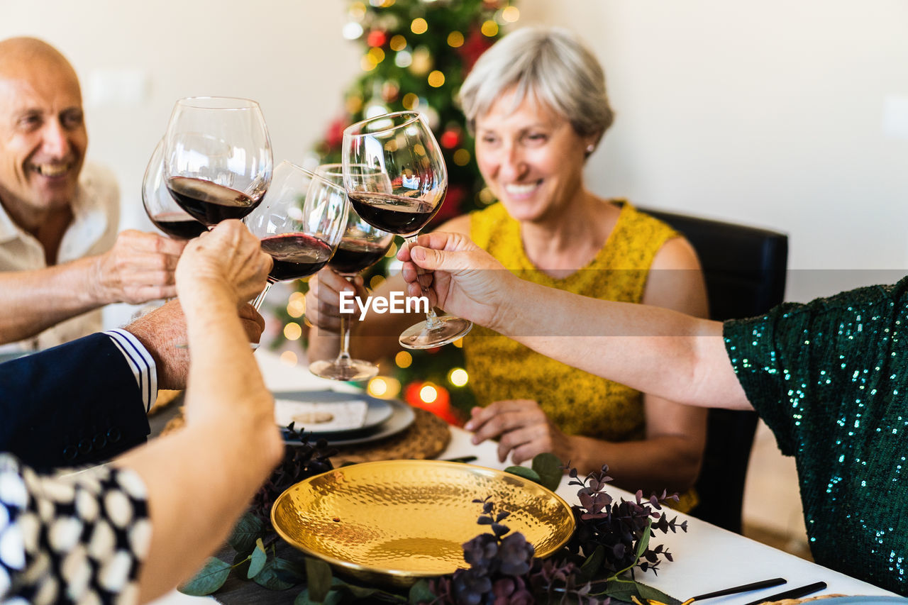 Happy mature friends gathering at festive table near shining christmas tree and clinking wineglasses while celebrating christmas eve together