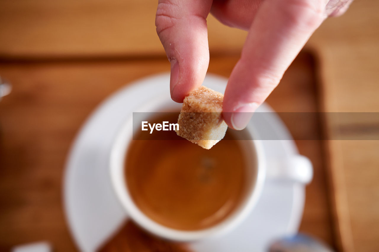 Cropped hand holding sugar cube over coffee cup on table