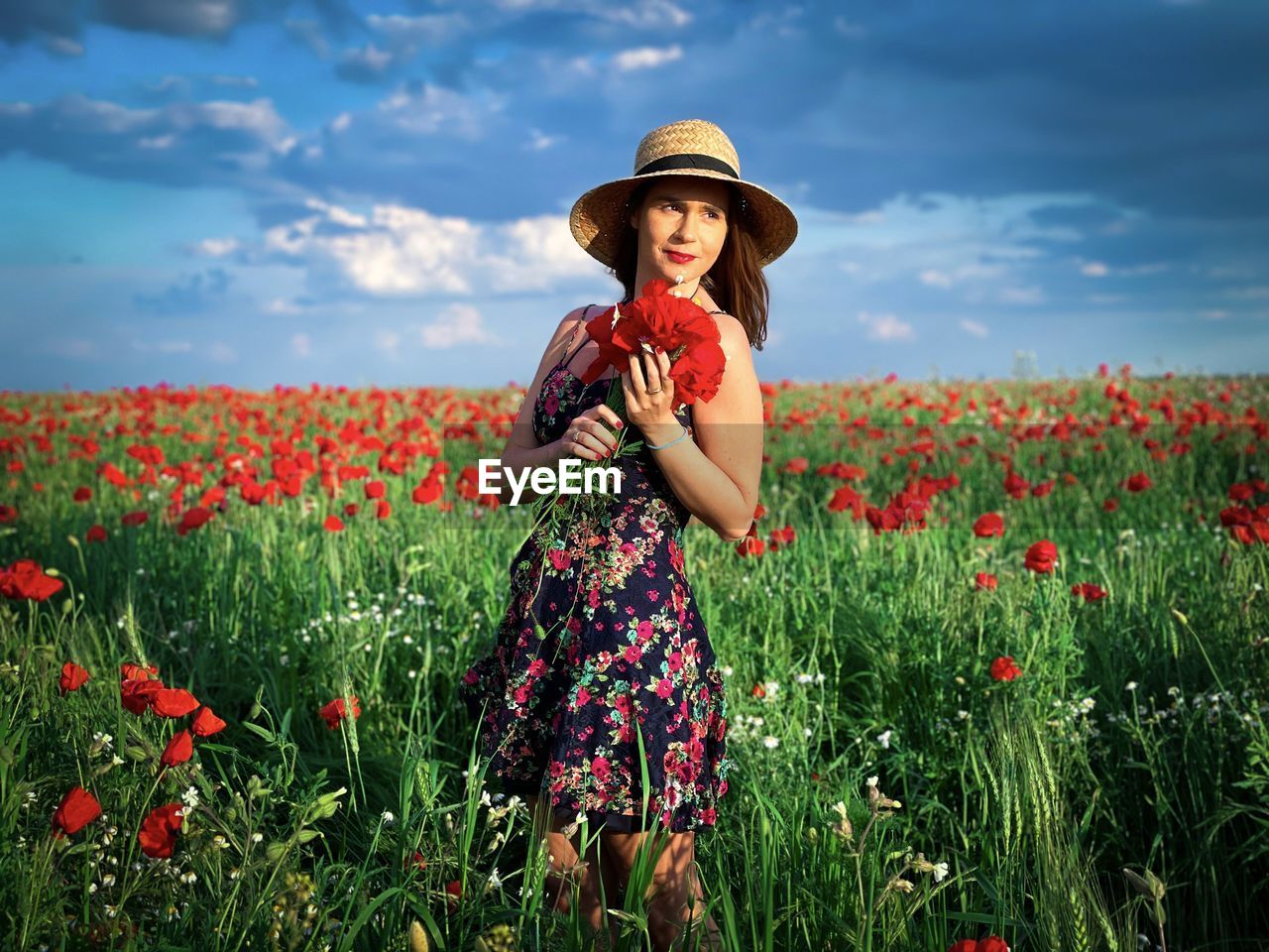 Woman wearing dress and hat holding a bouquet of poppies in a field of poppies