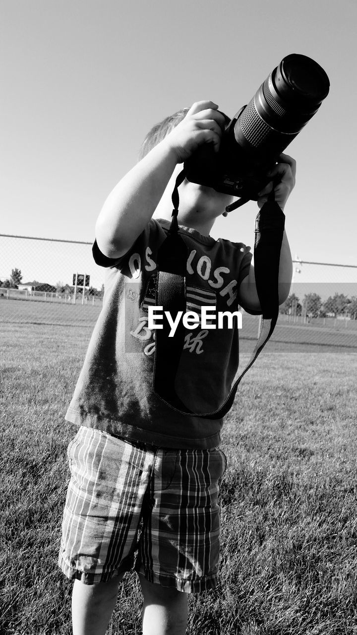Boy photographing with camera while standing on field against clear sky