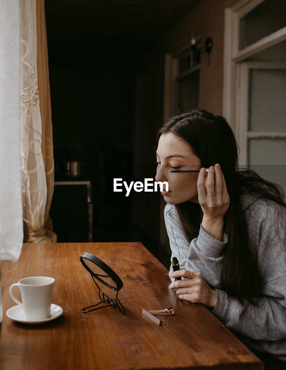 Attractive woman with the mirror doing makeup at home with cosmetic and coffee