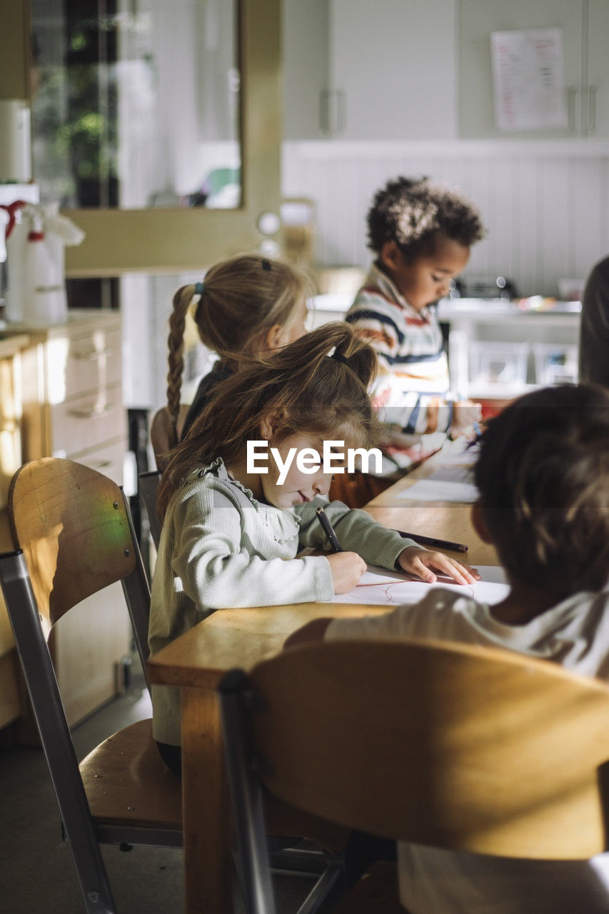 Girl writing on paper while sitting with students at bench in classroom