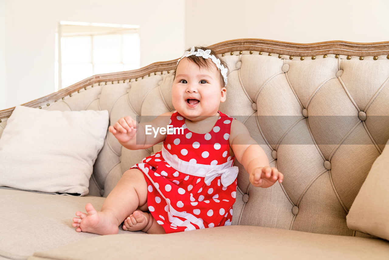 Happy asian cute baby girl in a red dress playing on the beige sofa at home