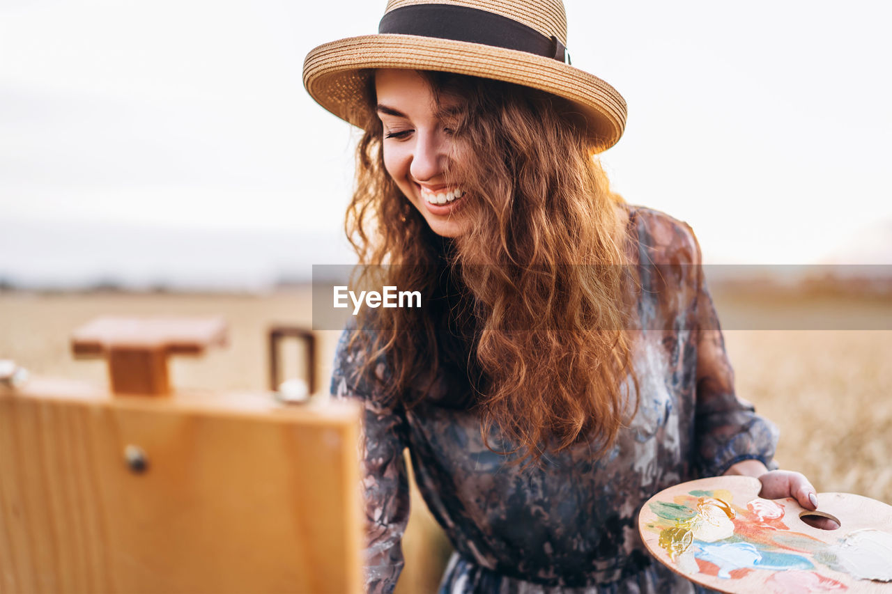 A young woman with curly hair and wearing a hat is painting in nature. 