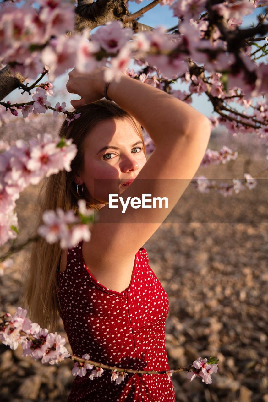 Blond woman with long hair posing under a flowering almond tree and looking at camera