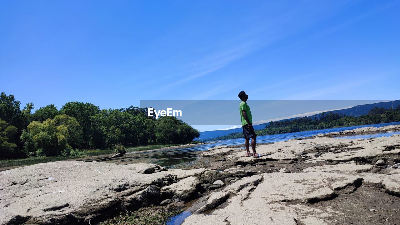sky, sea, one person, shore, coast, nature, water, land, full length, leisure activity, blue, beauty in nature, beach, scenics - nature, men, tree, lifestyles, body of water, clear sky, vacation, rock, day, holiday, ocean, rear view, trip, plant, adult, standing, activity, tranquility, outdoors, sunlight, environment, non-urban scene, landscape, sunny, tranquil scene, travel, casual clothing, travel destinations, mountain