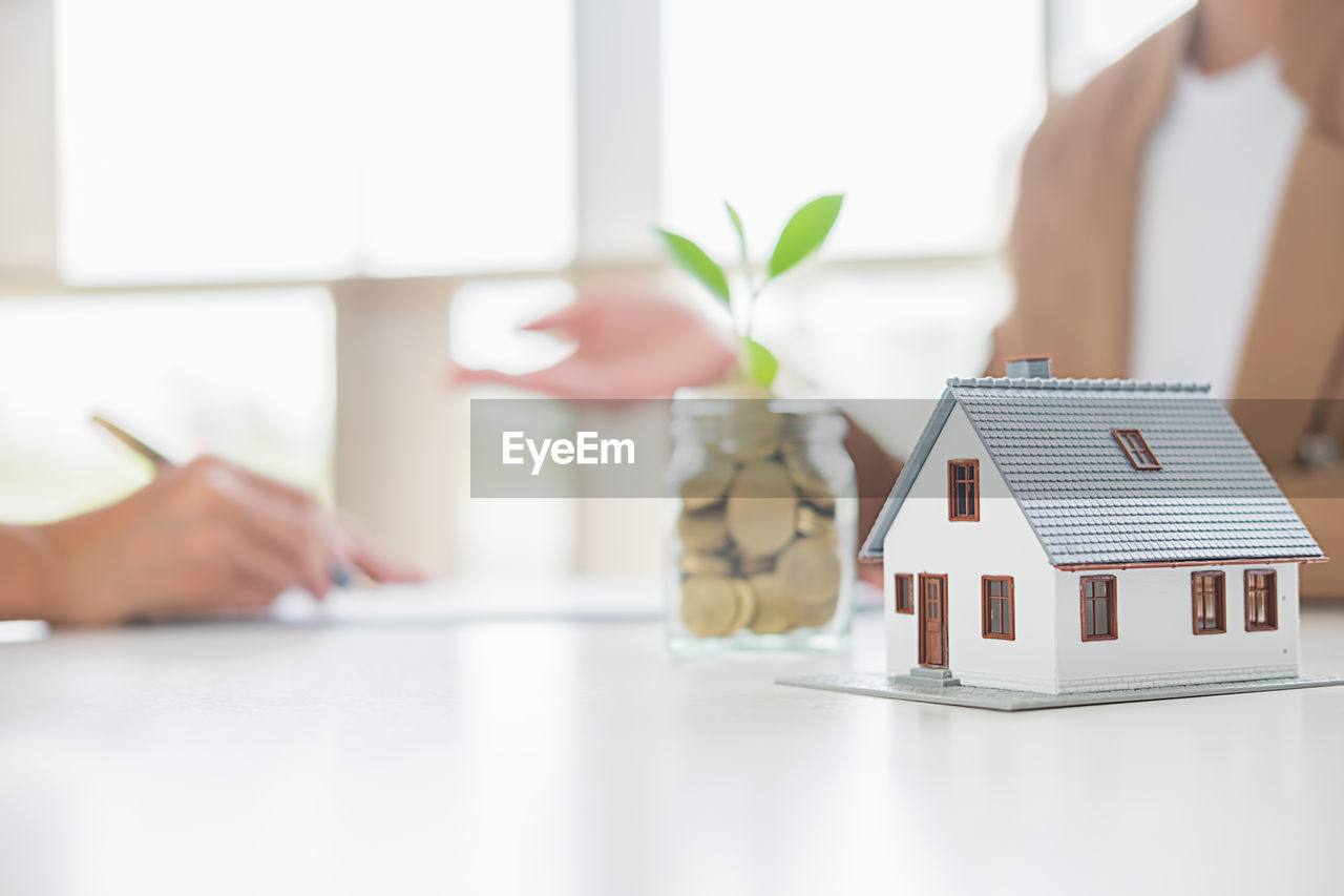 Midsection of real estate agent and client by model home and coins in jar on table 