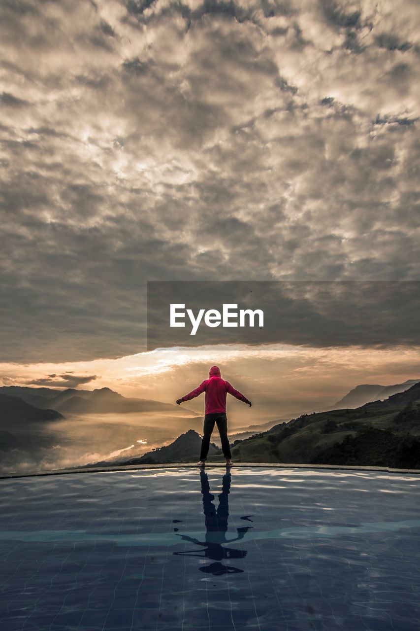 Man standing at infinity pool against cloudy sky during sunset