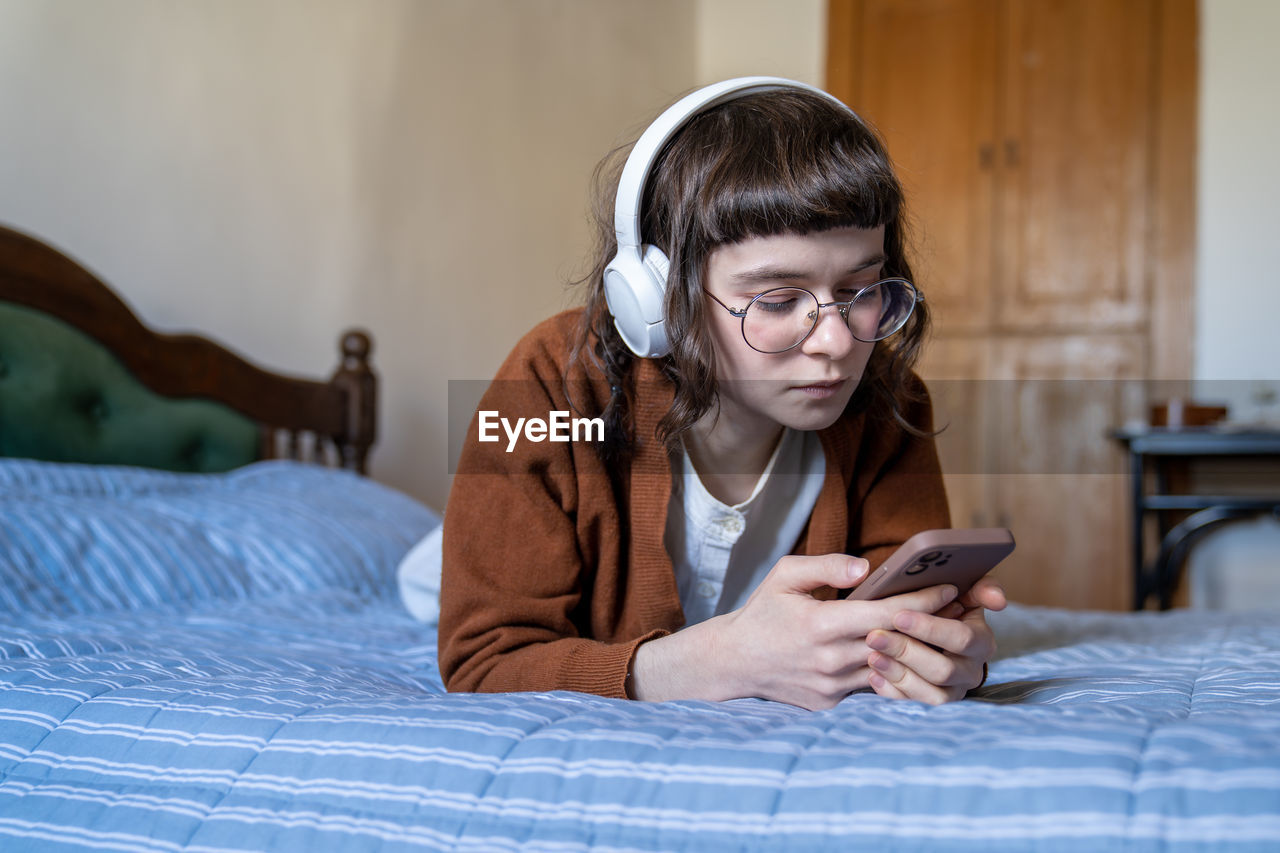 Relaxed music lover teenage girl listening to favourite single in headphones lying in bed at home.