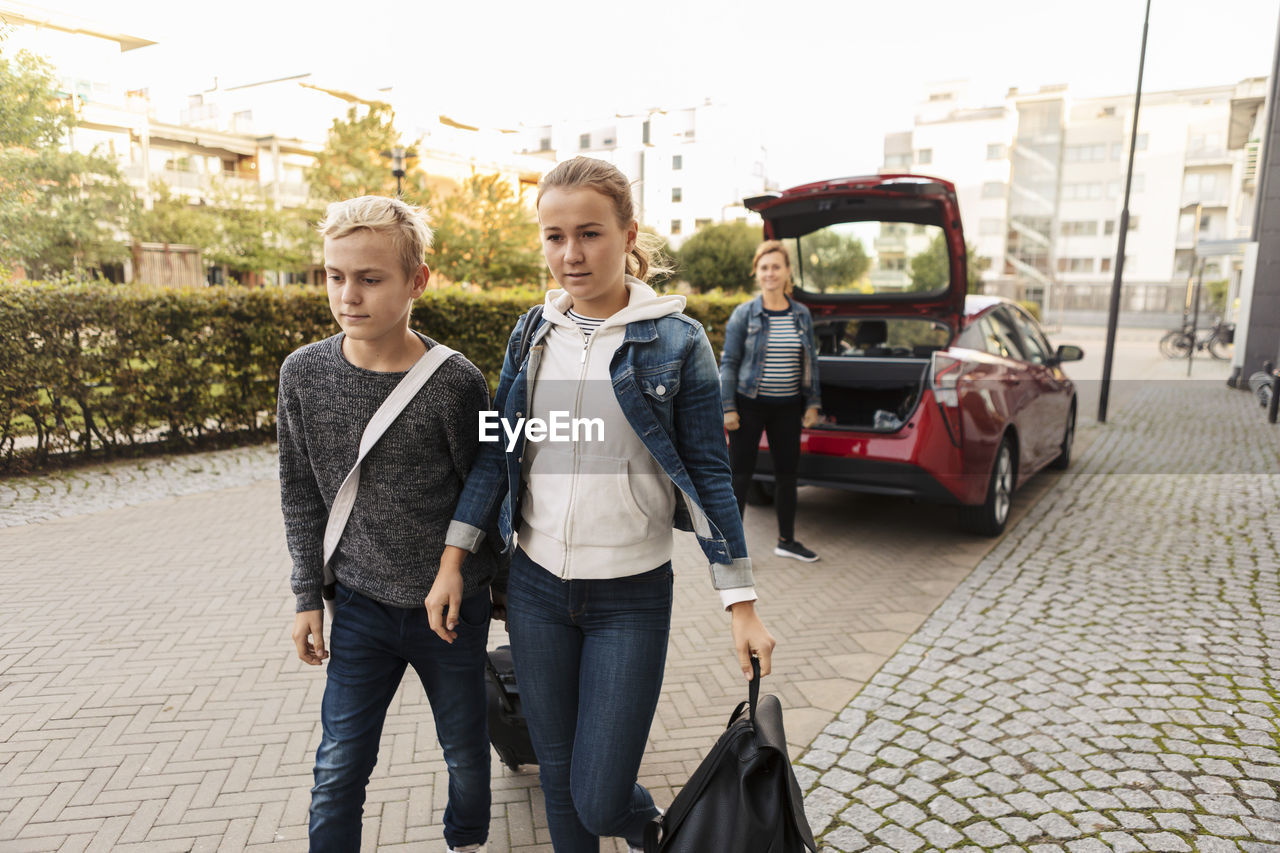 Siblings with suitcase walking while smiling mother standing by car on road