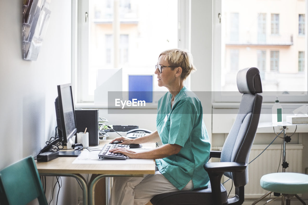 Side view of mature female doctor working over computer while sitting in clinic