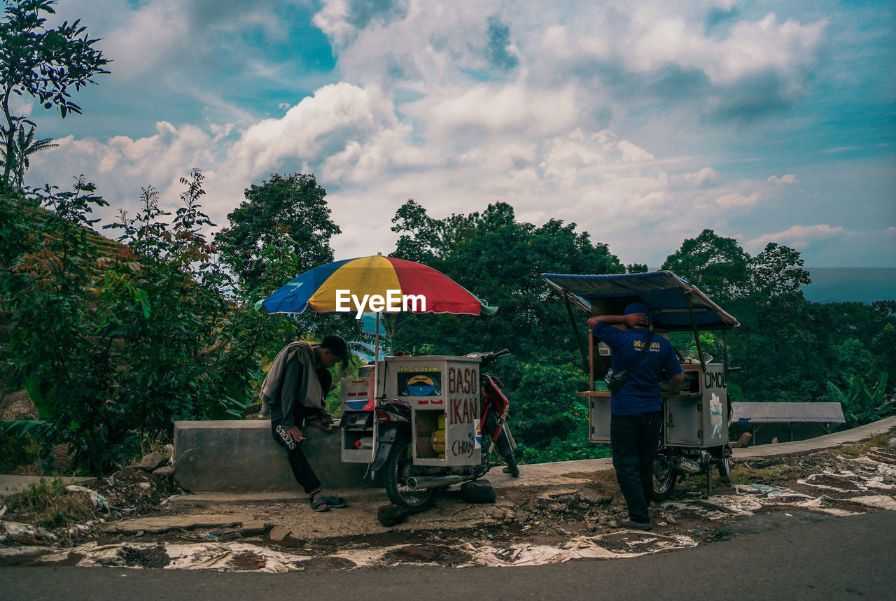 umbrella, cloud, sky, rural area, nature, transportation, tree, protection, plant, adult, rain, occupation, wet, men, environment, rickshaw, outdoors, land, landscape, road, beach, city, group of people, security, water, day, mode of transportation, asian style conical hat, rural scene, parasol, travel
