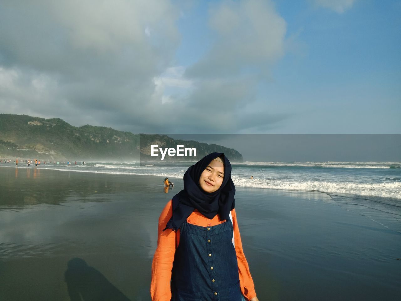 Portrait of young woman standing at beach against cloudy sky