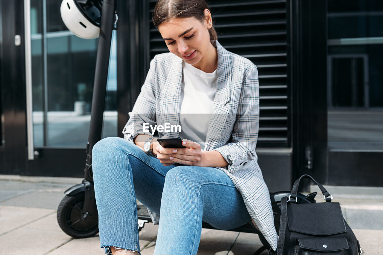 Businesswoman typing on a cell phone while sitting on electric push scooter at building	
