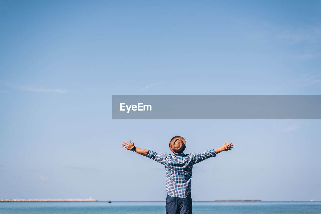 Rear view of man with arms outstretched standing by sea against sky