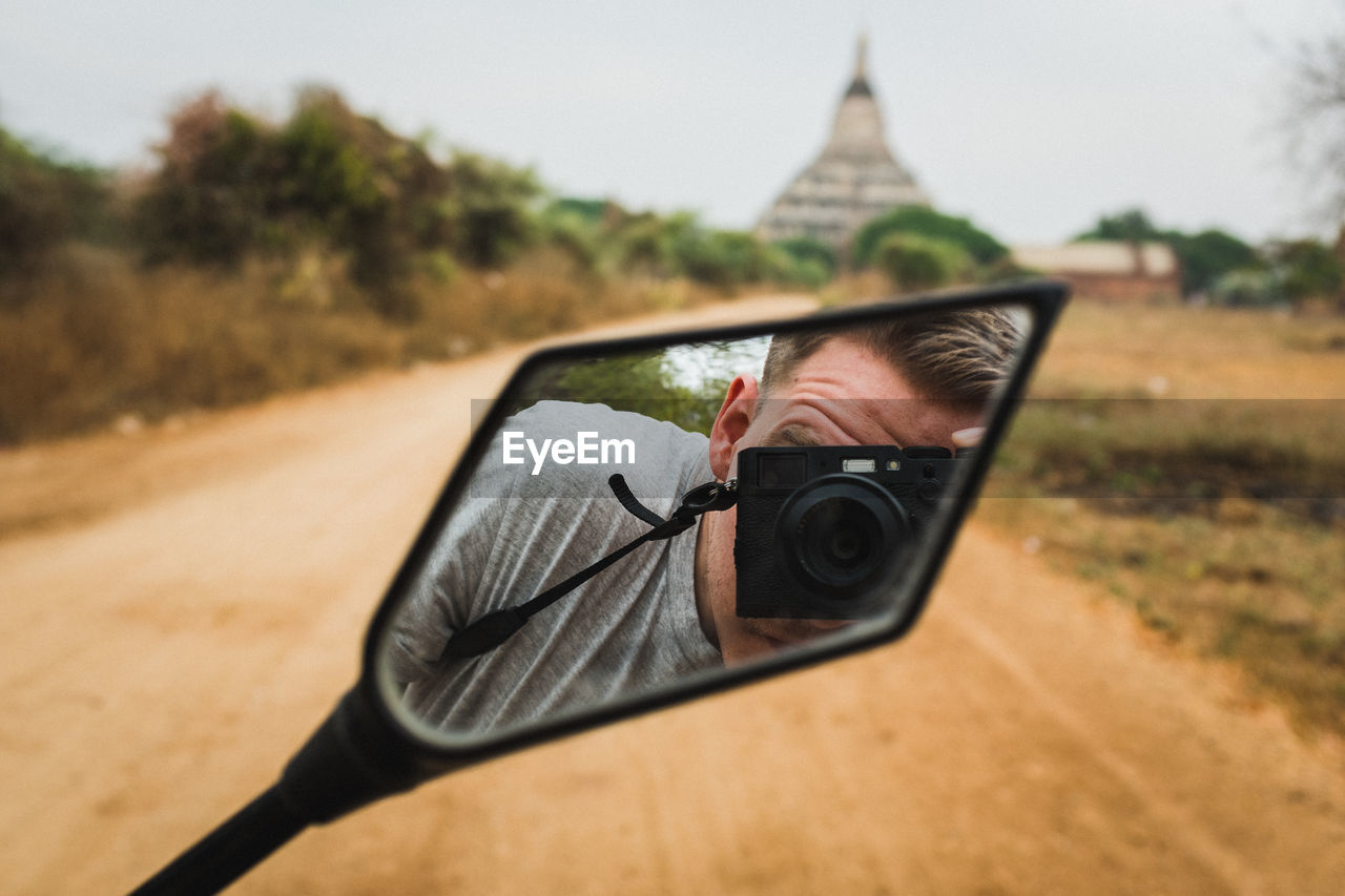 Reflection of man photographing with camera in side-view mirror of motorcycle on road