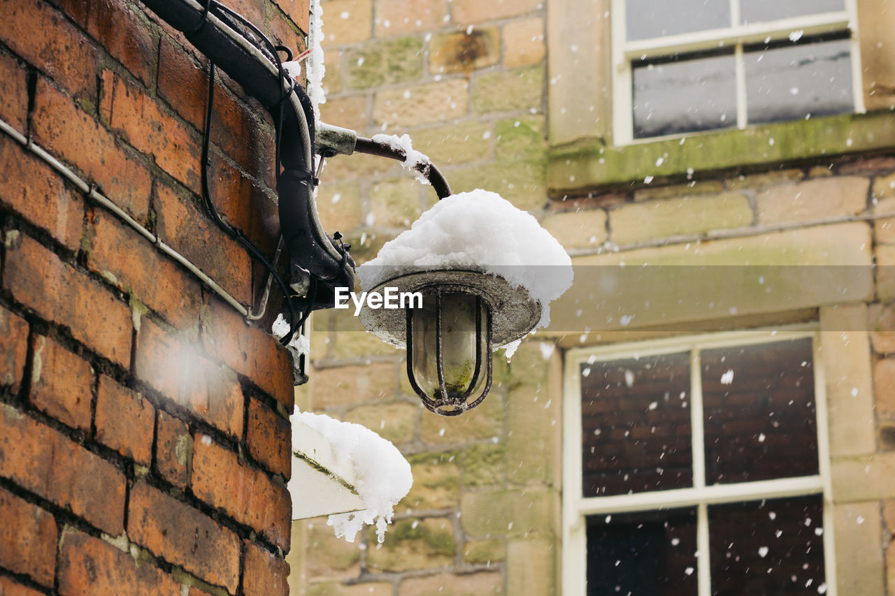 Low angle view of electric lamp mounted on brick wall during snowfall