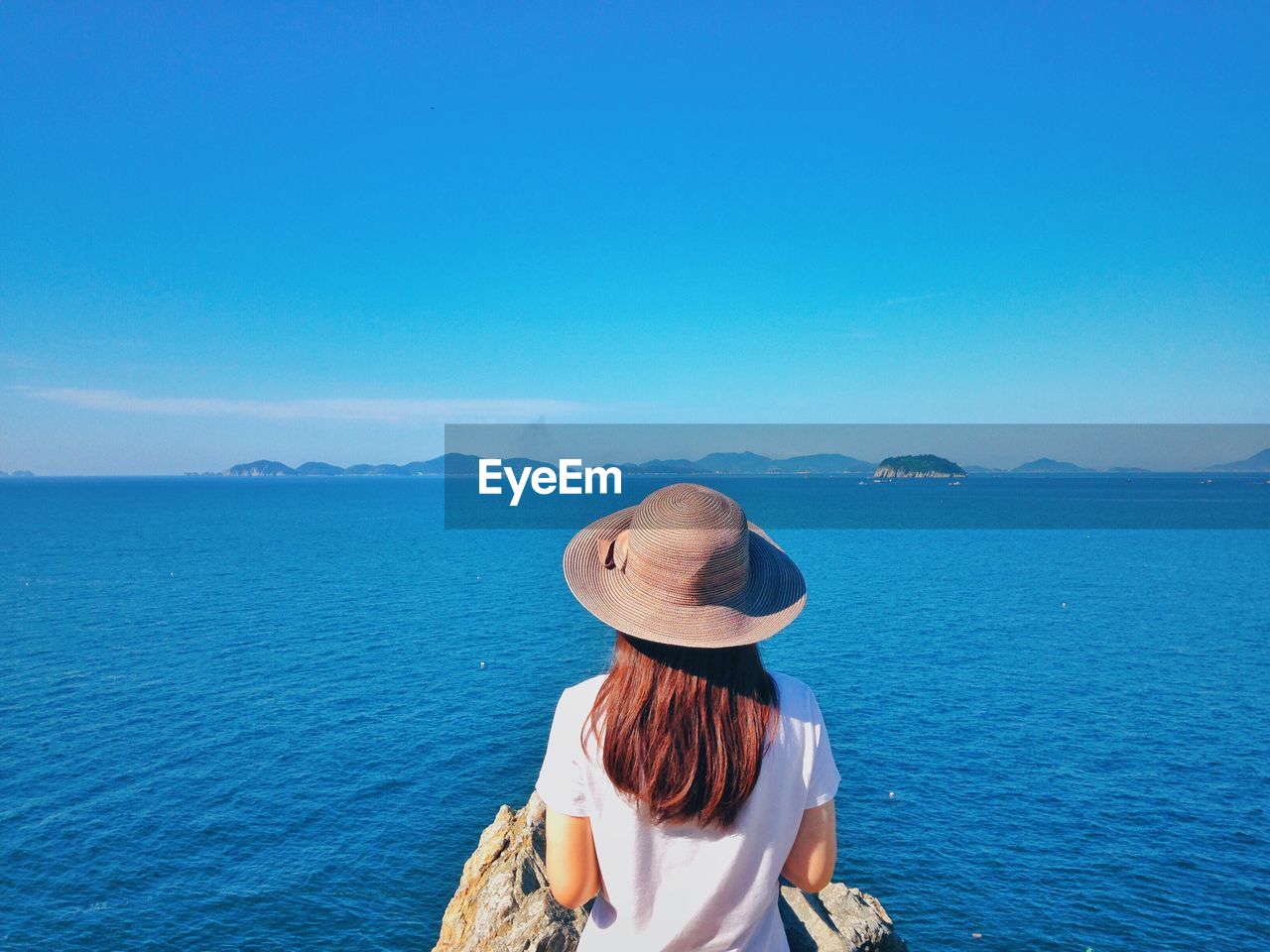 Rear view of woman looking at sea while standing against clear blue sky during sunny day