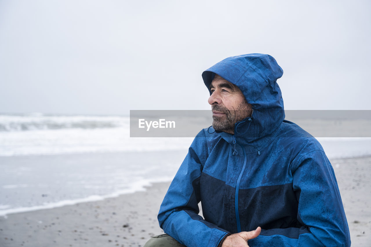 Contemplated mature man in blue raincoat crouching at beach against sky