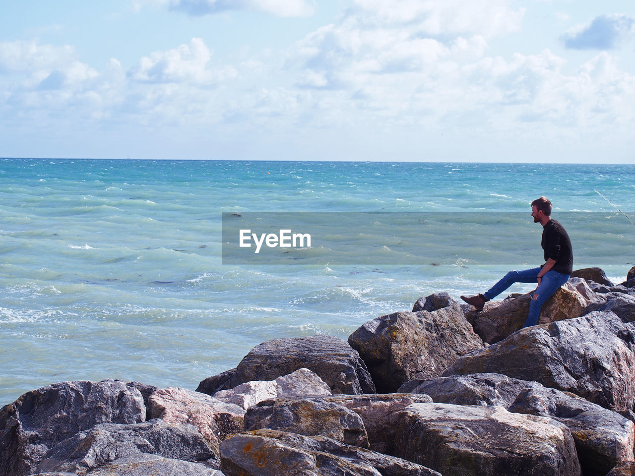 Side view of man looking at sea while sitting on rocks against sky