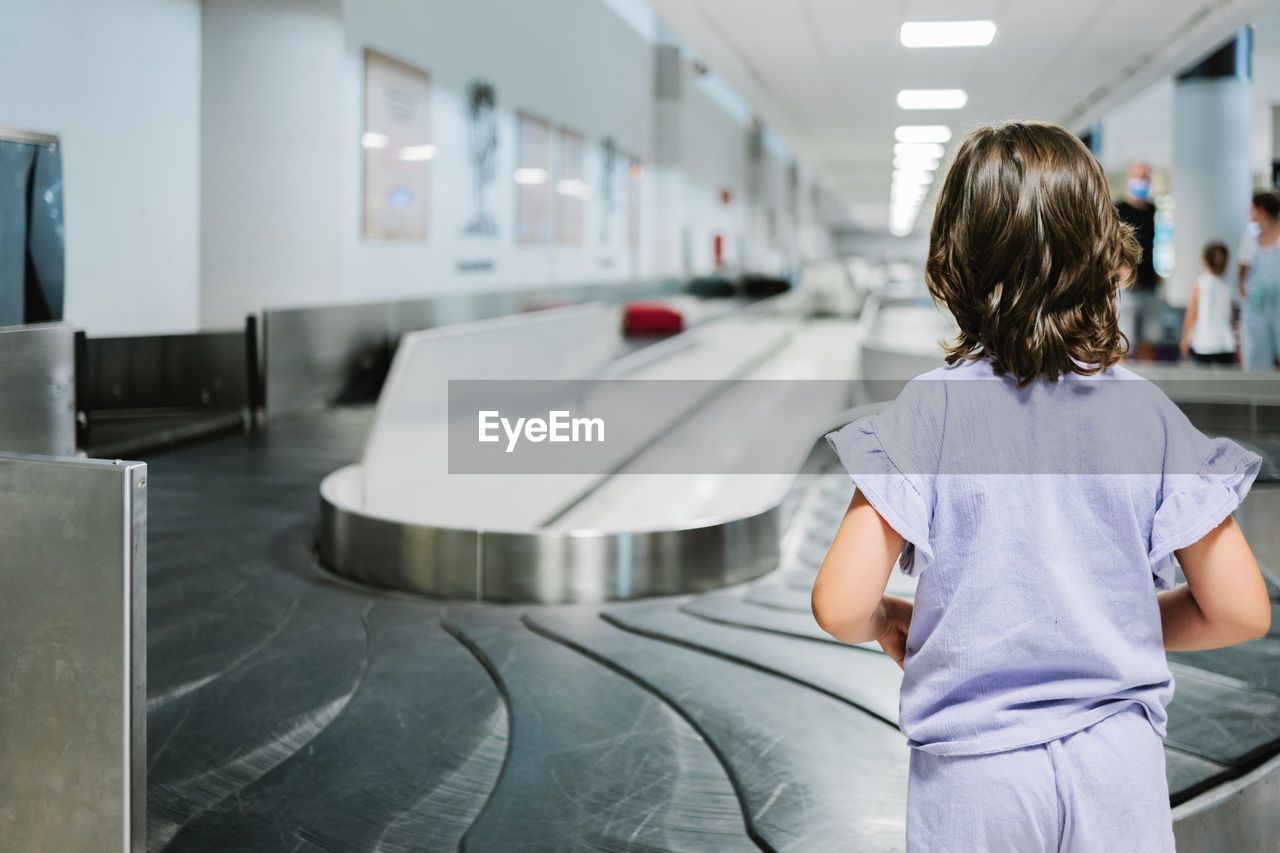 Back view of unrecognizable girl in lilac clothes waiting for luggage to arrive on carousel in airport terminal during trip