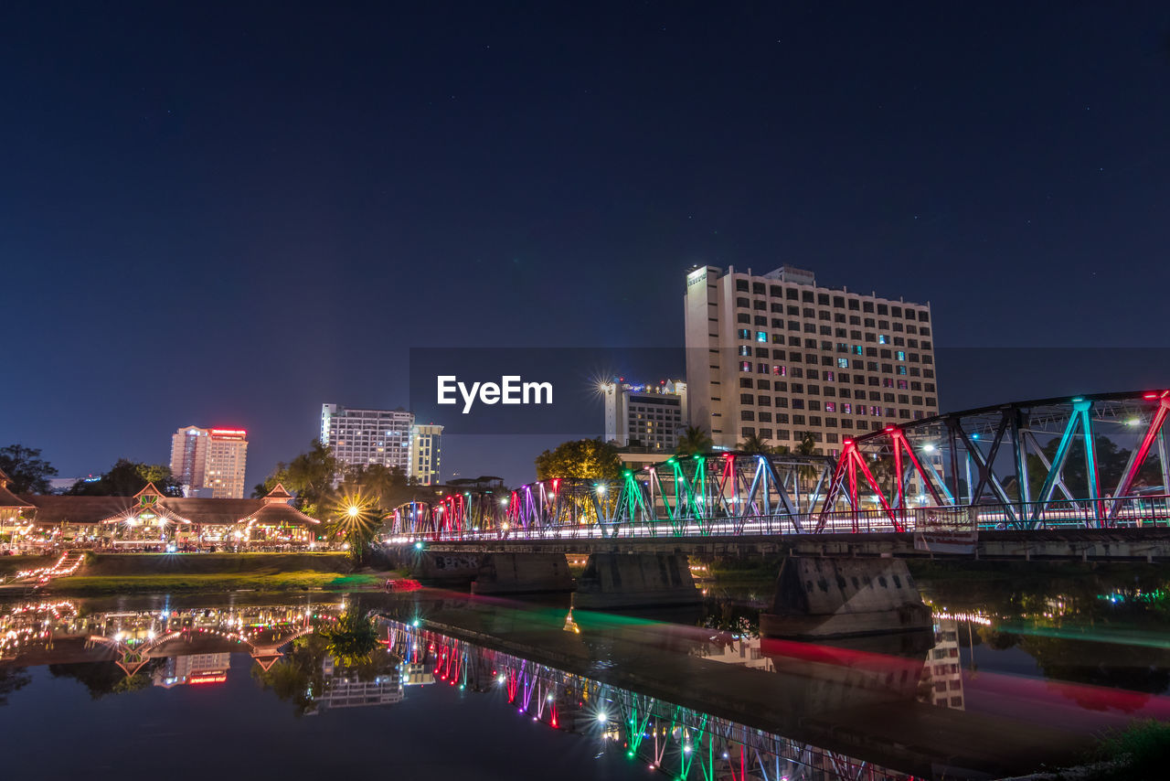 Illuminated modern buildings by river against sky at night