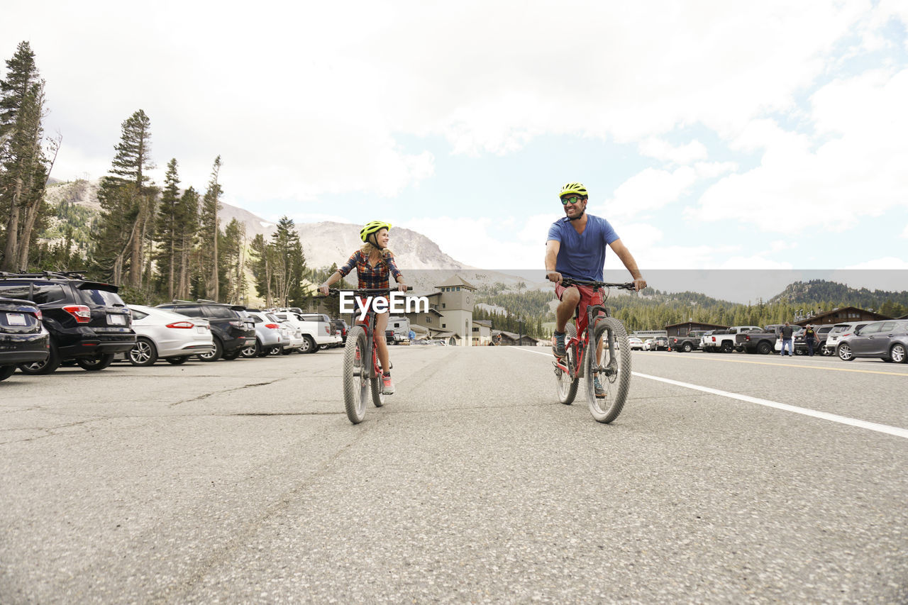 Happy couple riding bicycles on road against cloudy sky in forest