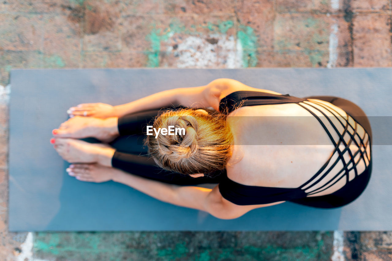 Full body from above of flexible female practicing paschimottanasana on sports mat on rooftop at sunset