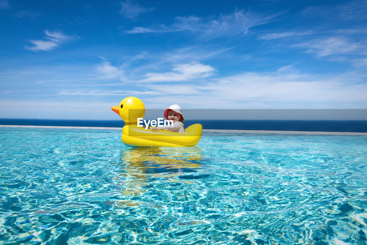 Baby girl with inflatable ring on infinity pool against blue sky