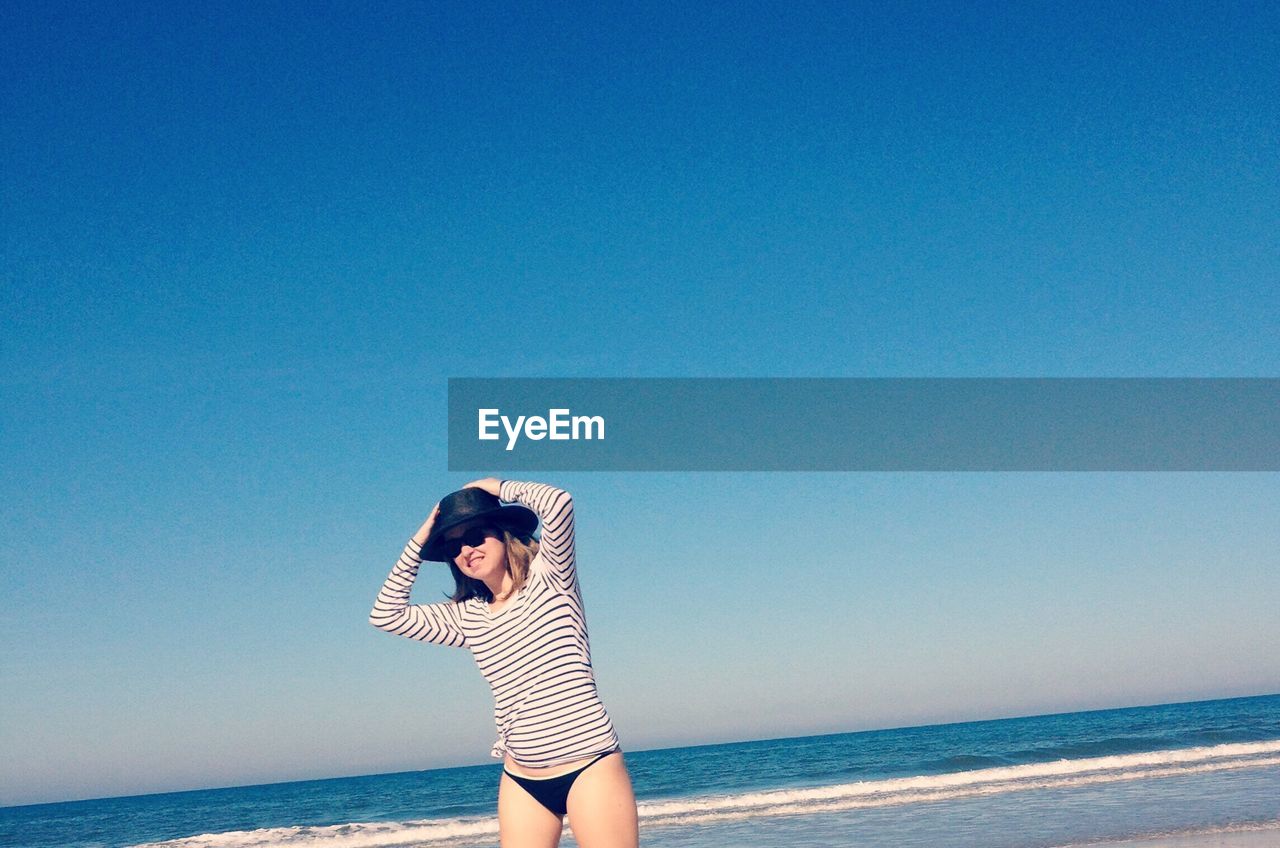 Young woman wearing hat and sunglasses at beach against clear blue sky on sunny day