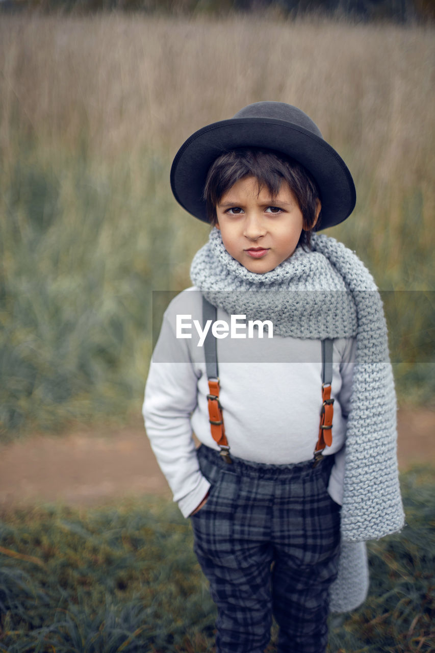 Boy child in plaid pants, hat, suspenders and scarf stands in a field in autumn