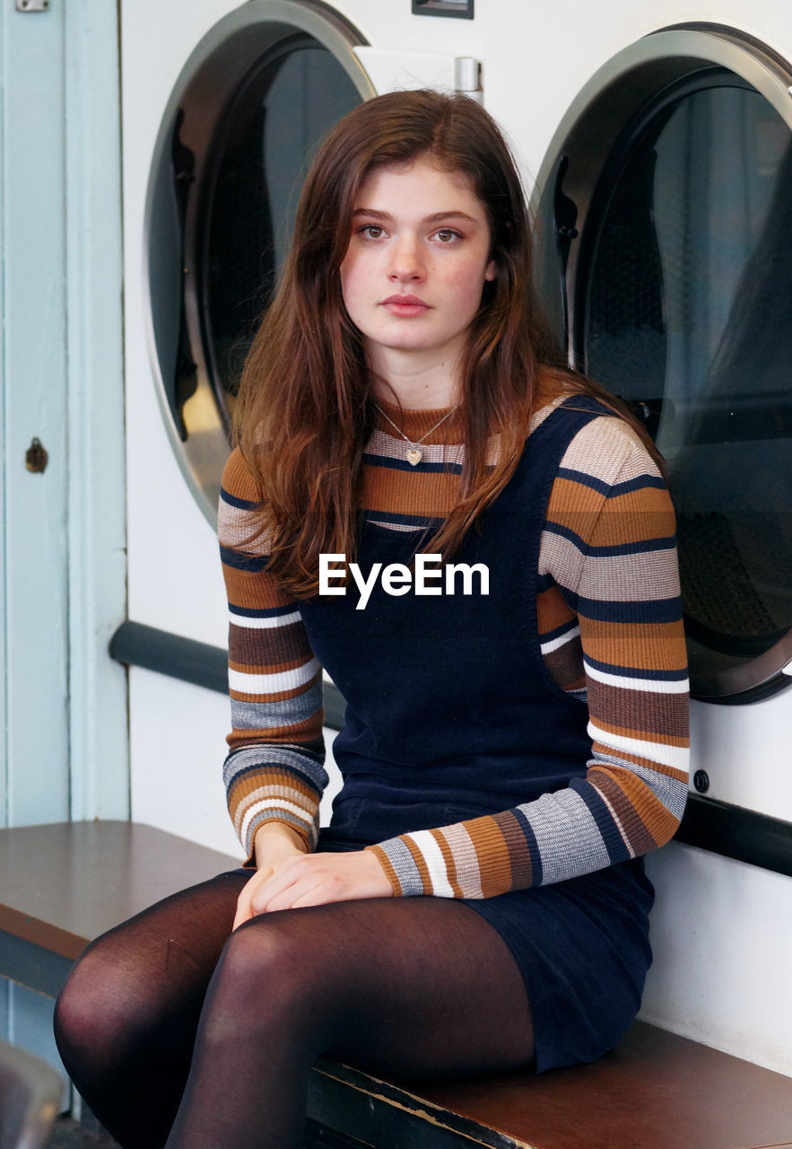 Portrait of beautiful young woman sitting against washing machine